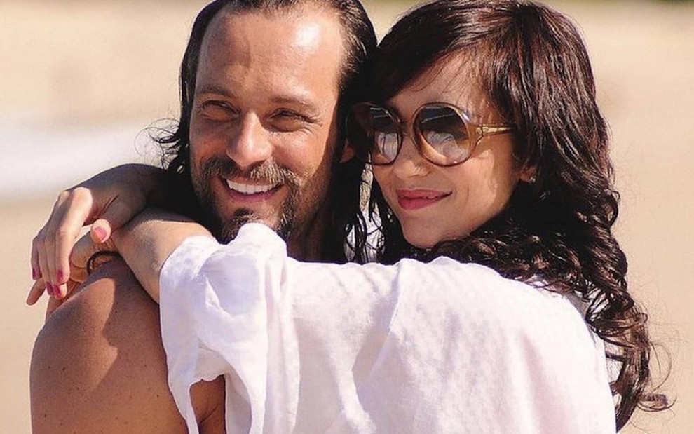 Sem camisa, Fernando Pavão posa sorrindo em uma praia com Simone Spoladore nas gravações de Pecado Mortal, da Record