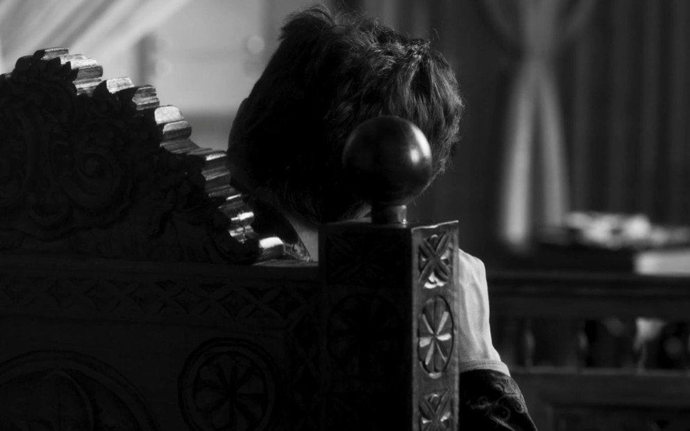 Um homem de costas sentado em um trono em uma cena em preto e branco da novela Reis