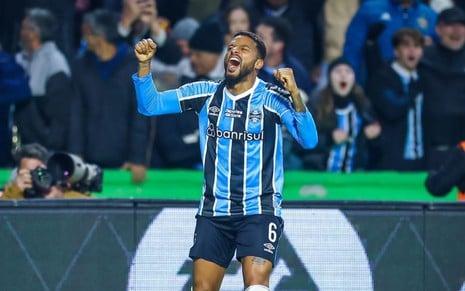 A imagem mostra um jogador de futebol durante uma comemoração em campo. Ele está vestindo o uniforme do Grêmio, que consiste em uma camisa listrada verticalmente nas cores azul, preto e branco, com o número 6 visível no calção preto