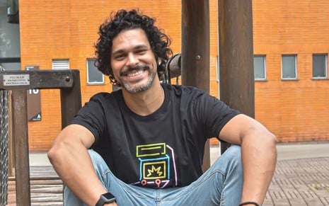 De camiseta azul e calça jeans, Renan Monteiro está sentado com os braços apoiados na perna