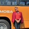 Na imagem, uma pessoa está encostada em um ônibus amarelo com a inscrição "Viação Formosa". Ela usa óculos, um blazer vermelho e uma camisa estampada.