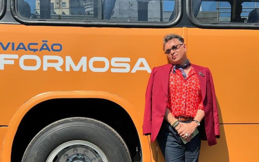 Na imagem, uma pessoa está encostada em um ônibus amarelo com a inscrição "Viação Formosa". Ela usa óculos, um blazer vermelho e uma camisa estampada.