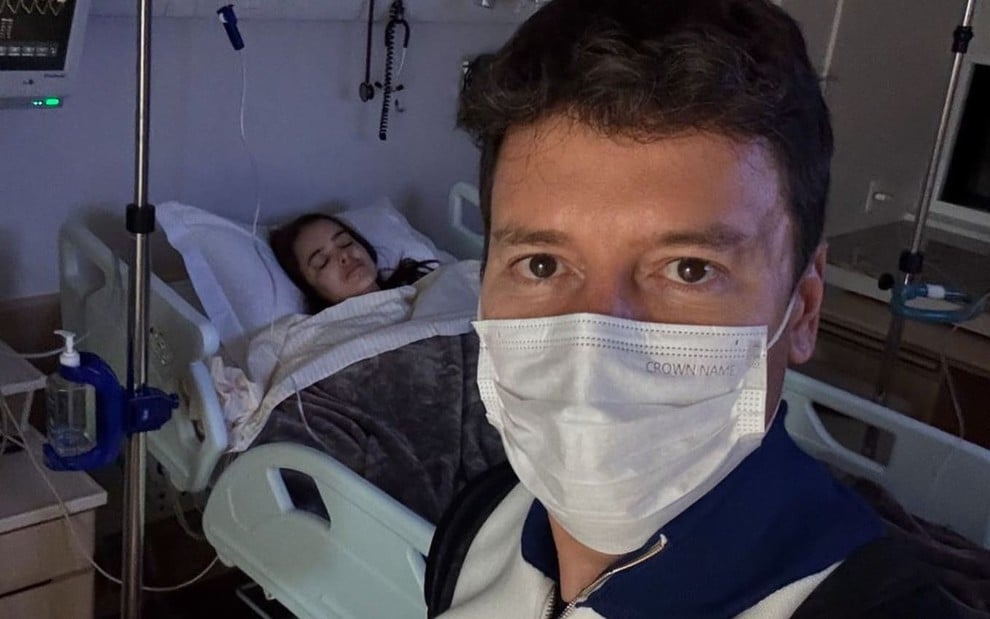 Rodrigo Faro usa máscara e está na frente de um leito hospitalar, no qual Vera Viel está deitada