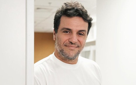 Rodrigo Lombardi usa uma camiseta branca e sorri discretamente, enquanto apoia o corpo no batente de uma porta
