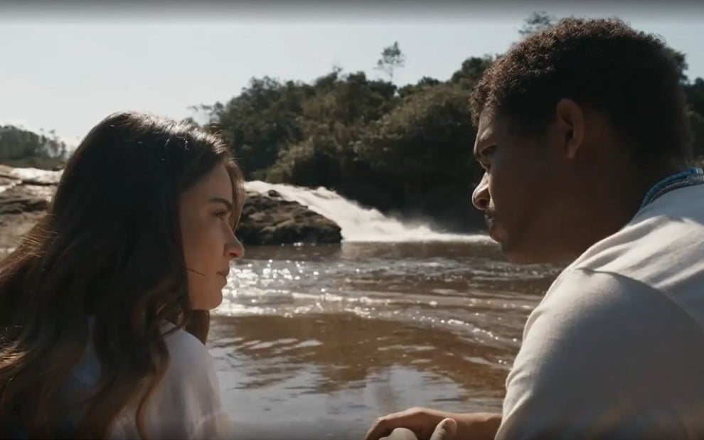 Em cena de Renascer, Giullia Buscacio e Juan Paiva conversam com uma cachoeira ao fundo