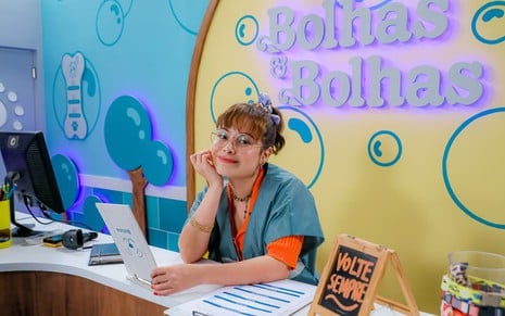 Thay Bergamim usa uma blusa laranja e um avental azul; ela está apoiada sobre uma mesa, encara a câmera e dá um leve sorriso. Ao fundo, é possível ver o logo "Bolhas & Bolhas"