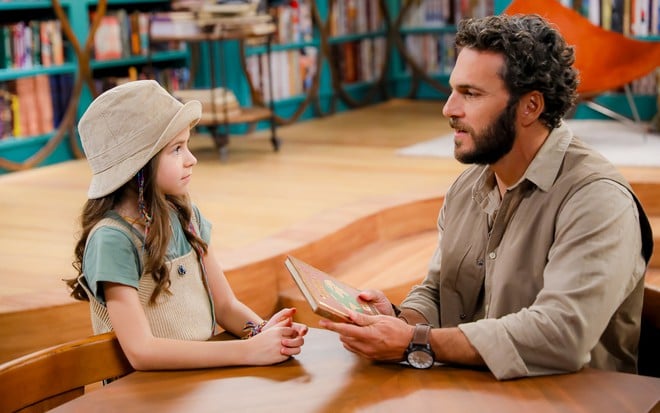 Anna (Mel Summers) sentada em uma mesa em frente a Paulo (Elam Lima) em cena de A Caverna Encantada