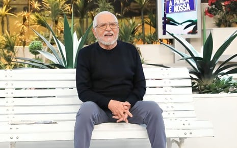 Carlos Alberto de Nóbrega está sentado no banco de A Praça É Nossa, com uma blusa preta e calça jeans