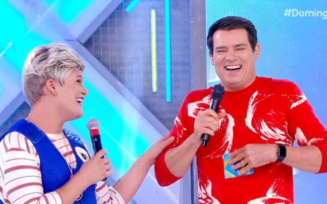 Maicon Sales, vestido como sua personagem Ana Maria Brisa, gargalha ao lado de Celso Portiolli no cenário do Domingo Legal