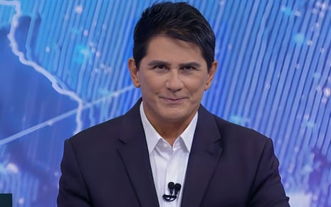 A imagem mostra um apresentador de notícias com cabelo escuro e curto, usando terno escuro e camisa branca. Ele sorri levemente, com fundo azul.