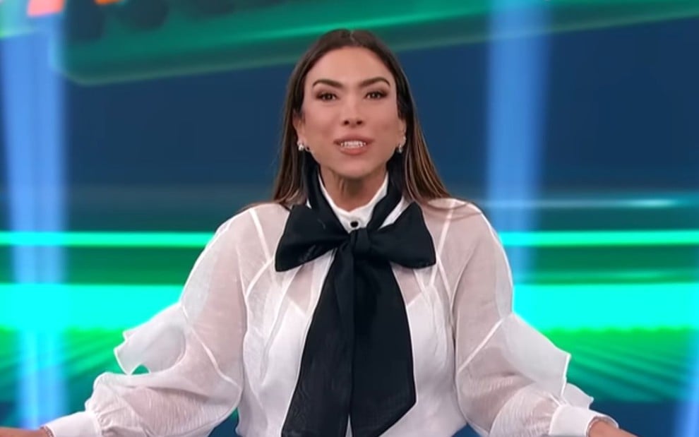 A imagem mostra uma apresentadora de TV em um estúdio, usando uma blusa branca transparente com uma grande laço preto na altura do pescoço.