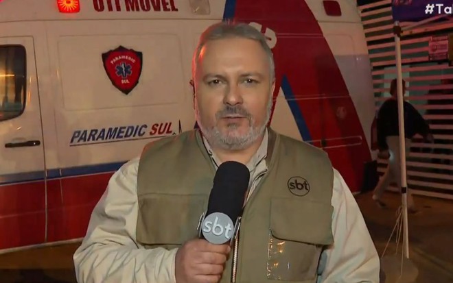 Emerson Tchalian com um colete com o símbolo do SBT e um microfone com o logo da emissora