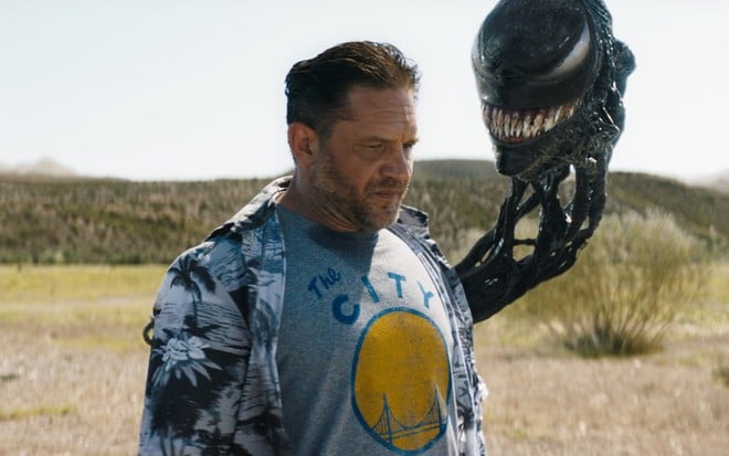 Com roupa de turista americano em férias, Tom Hardy parece triste e é observado por uma cabeça gosmenta de Venom