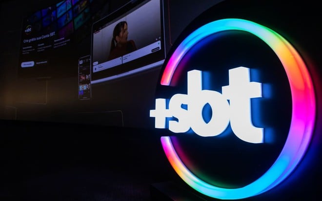 Logo do +SBT em gesso e com iluminação de LED; ao fundo, é possível ver uma projeção da interface da plataforma