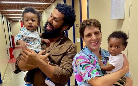 Amaury Lorenzo segura e olha para um bebê; Diego Martins segura um outro bebê e sorri para a câmera em cena de Terra e Paixão