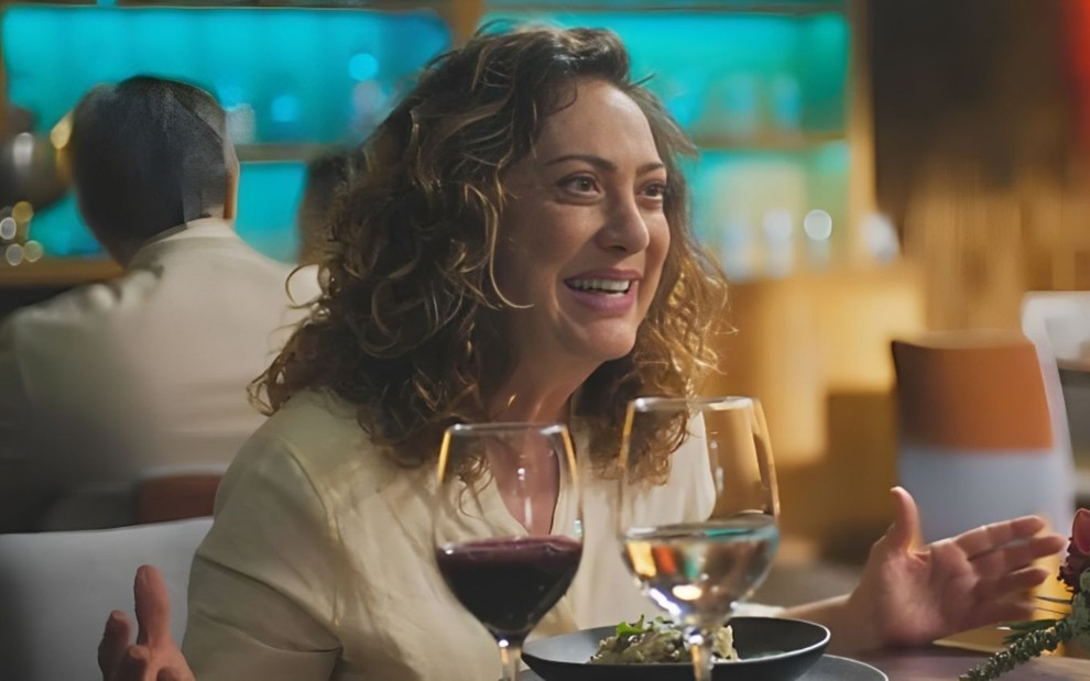 A atriz Eliane Giardini sorrindo, em cena em restaurante, com taças de vinho e de água sua em frente, em Terra e Paixão