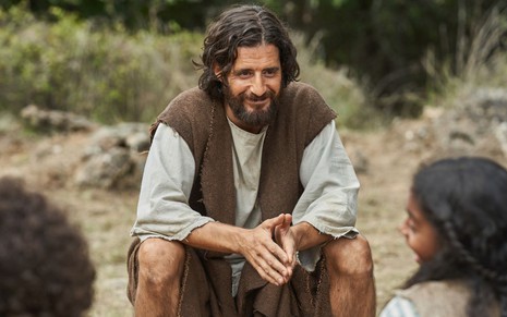 Jonathan Roumie está caracterizado como Jesus Cristo; sentado em um tronco, ele sorri para crianças (que estão de costas)