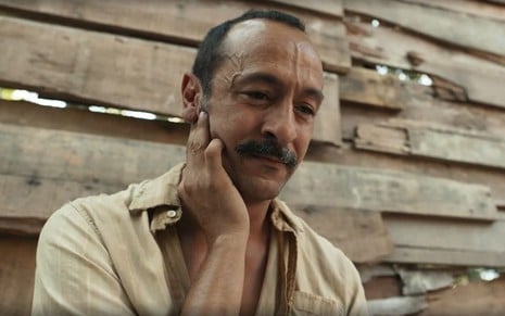 Em cena de Renascer, Irandhir Santos está olhando para frente, com a mão no rosto, pensativo; ele usa bigode