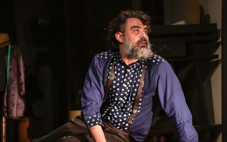 Com barba espessa, Carmo Dalla Vecchia está sentado no chão e tem expressão de desespero