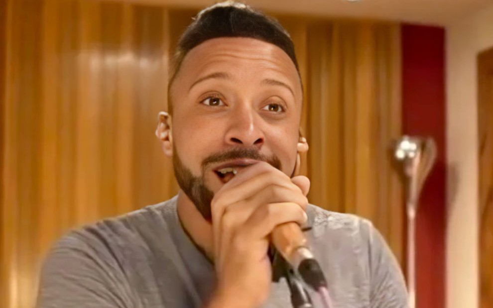 O cantor sertanejo Ton Ferreira segura um microfone; ele está com uma camisa cinza em um estúdio