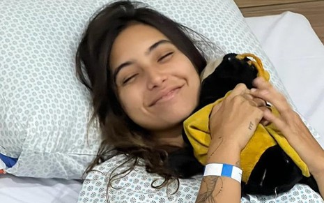 Vanessa Lopes está deitada em uma cama de hospital, abraçada com a pelúcia de uma abelha