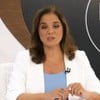 Uma mulher com cabelos lisos, até os ombros, de blazer branco e blusa azul está com expressão séria em uma bancada de um programa de entrevistas