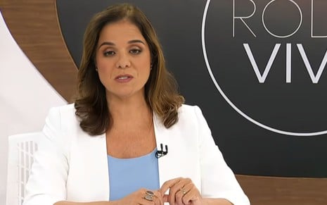 Uma mulher com cabelos lisos, até os ombros, de blazer branco e blusa azul está com expressão séria em uma bancada de um programa de entrevistas