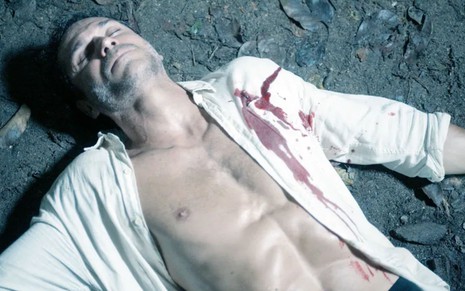 Paulo Rocha caído no chão desacordado e com mancha de sangue na camisa branca em cena da novela Terra e Paixão