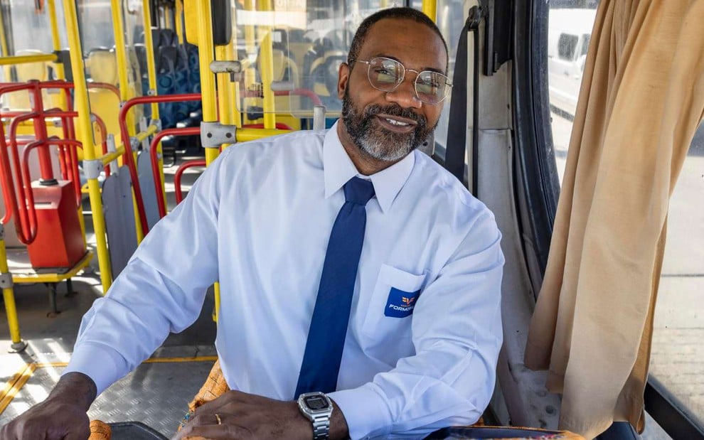 O ator MV Bill sorrindo em cena como Lindomar, dentro de ônibus, em Volta por Cima