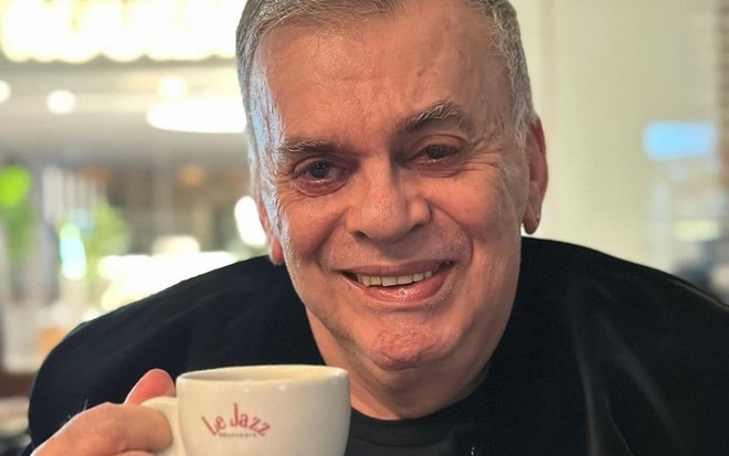 Walcyr Carrasco usa um terno preto e segura uma xícara de café branca; ele sorri enquanto encara a câmera