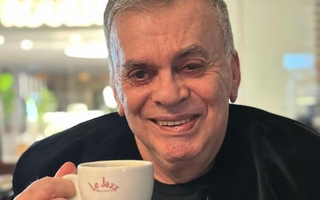 Walcyr Carrasco usa um terno preto e segura uma xícara de café branca; ele sorri enquanto encara a câmera