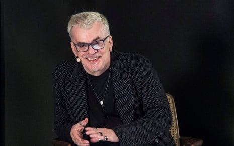 Foto do autor Walcyr Carrasco, de preto, em uma cadeira, com o fundo rpeto