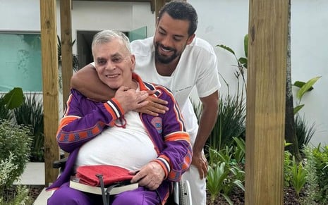 Sentado em uma cadeira de rodas, Walcyr Carrasco ganha um abraço do médico Pedro Andrade