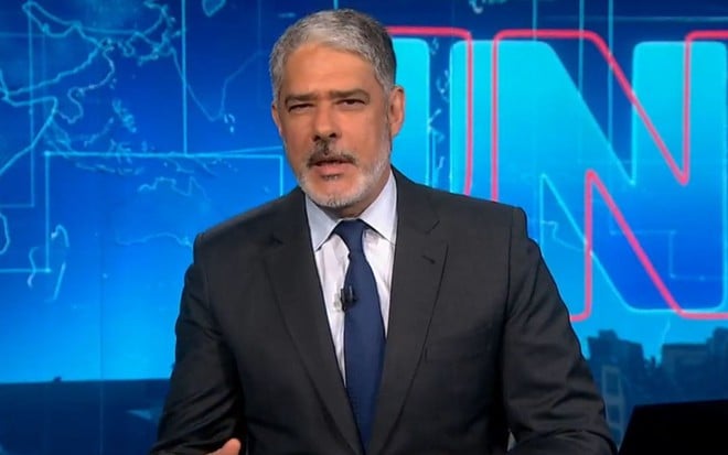 William Bonner está na bancada do Jornal Nacional; ele usa terno preto, gravata azul e está com expressão séria