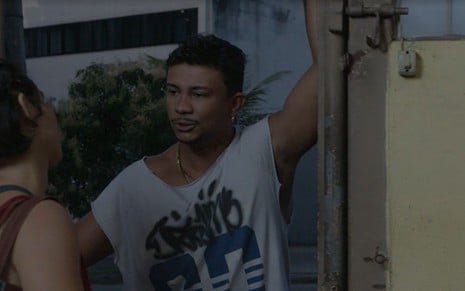 Em cena de Justiça, Xamã está parado no batente de uma porta, falando com Nanda Costa, que está de costas
