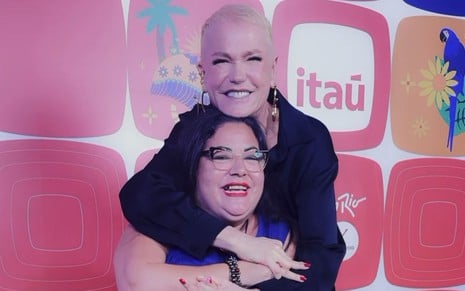 Xuxa Meneghel abraça Patrícia Veloso Martins por trás; elas estão em frente a painel de patrocinadores do Rock in Rio