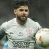 Na imagem, um jogador de futebol com barba e cabelo curto e molhado, usando a camisa cinza do Corinthians, está em ação próximo de uma bola.