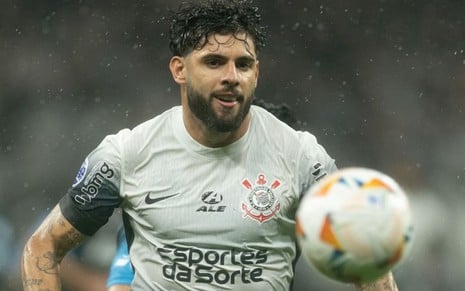 Na imagem, um jogador de futebol com barba e cabelo curto e molhado, usando a camisa cinza do Corinthians, está em ação próximo de uma bola.