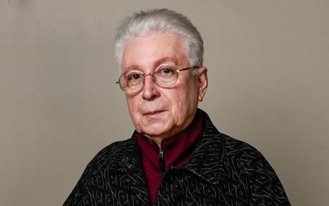 Homem branco, de cabelos grisalhos e curtos, usando camisa vermelha e casaco preto.