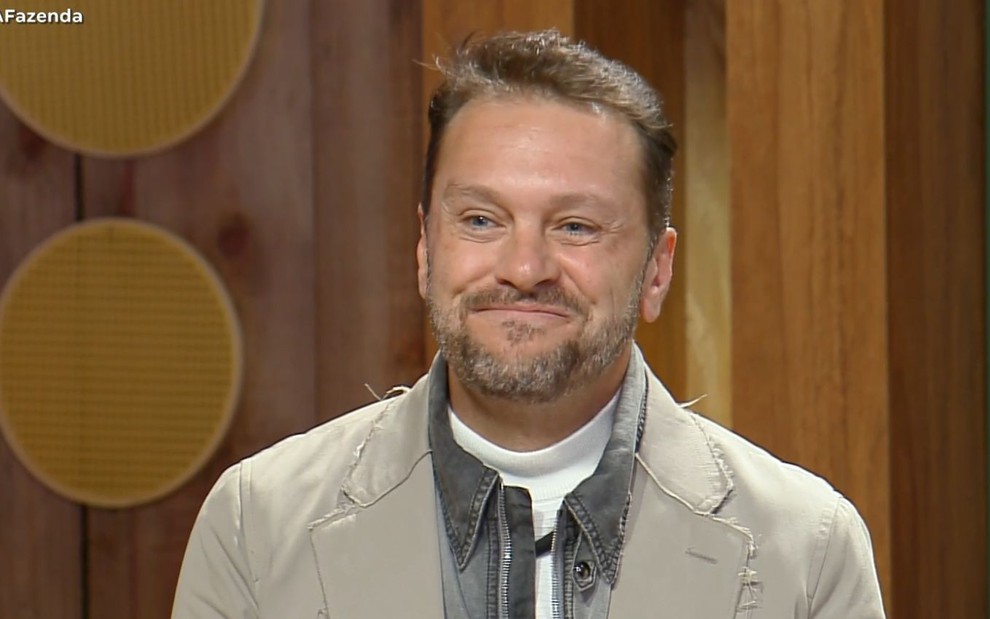 Um homem com barba e cabelo curto sorri suavemente em um cenário de madeira. Ele veste blazer bege e gola alta branca.