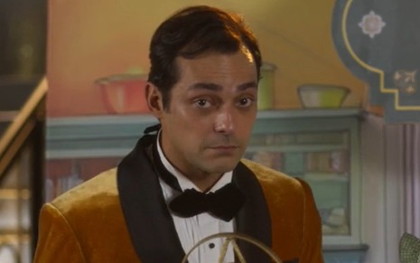 De terno, Eduardo Sterblitch fala em microfone antigo em cena da novela Garota do Momento