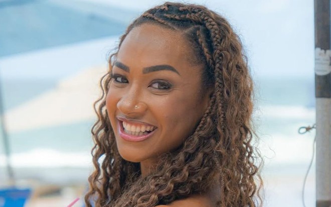 Aline Patriarca está sorrindo, segurando um coco na praia