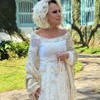 Ana Maria Braga usa vestido de noiva off-white com detalhes rendados e mangas bordadas. Ela segura um terço nas mãos e tem uma flor no cabelo.