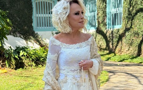 Ana Maria Braga usa vestido de noiva off-white com detalhes rendados e mangas bordadas. Ela segura um terço nas mãos e tem uma flor no cabelo.