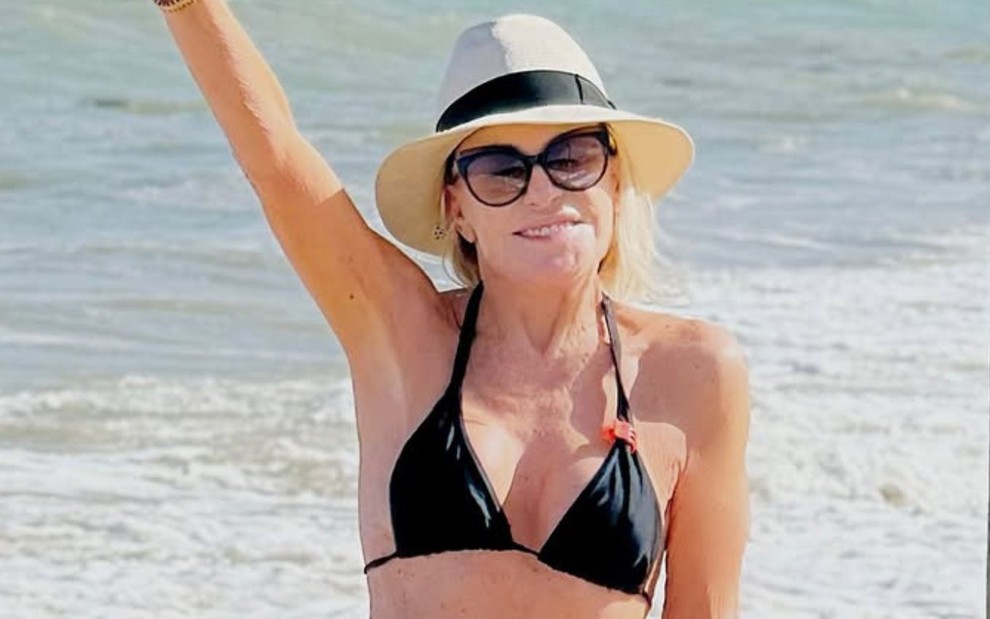 Ana Maria Braga está de biquíni e óculos de sol em uma praia, e estende a mão direita para cima