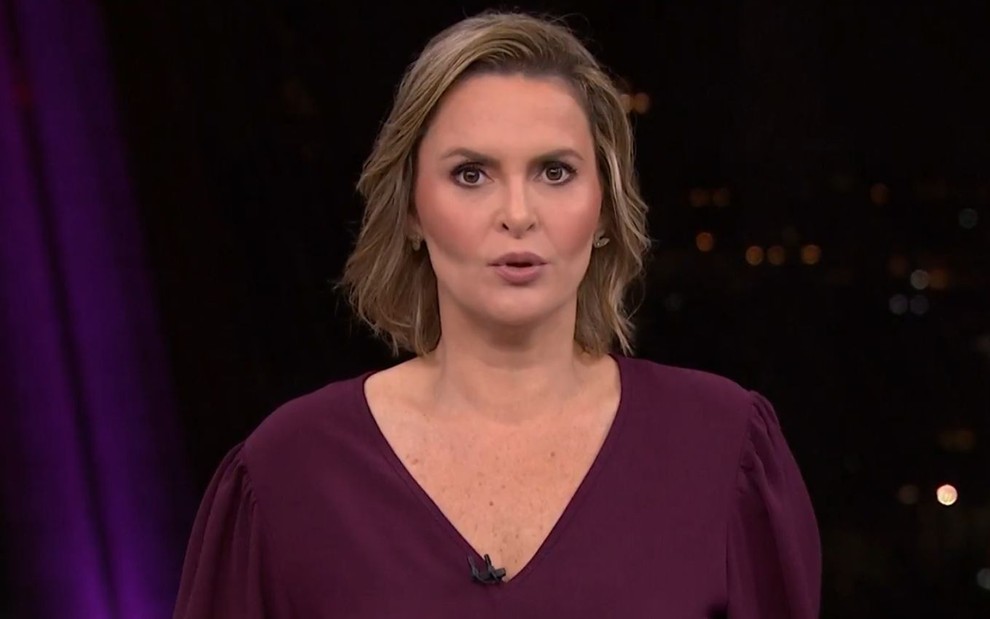 Mulher branca de cabelo loiro curto aparece em estúdio, usa blusa roxa e olha séria para a câmera.