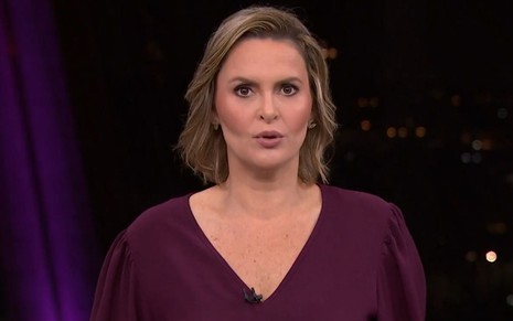 Mulher branca de cabelo loiro curto aparece em estúdio, usa blusa roxa e olha séria para a câmera.