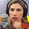 Ana Paula Minerato está com um fone de ouvido, na frente de um microfone com a logo da Band FM; ela está séria