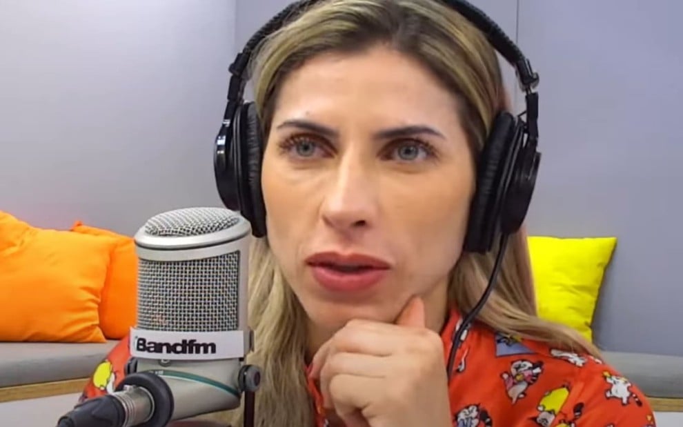 Ana Paula Minerato está com um fone de ouvido, na frente de um microfone com a logo da Band FM; ela está séria