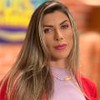 Ana Paula Minerato usa uma blusa lilás e um casaco laranja; ela encara a câmera, séria